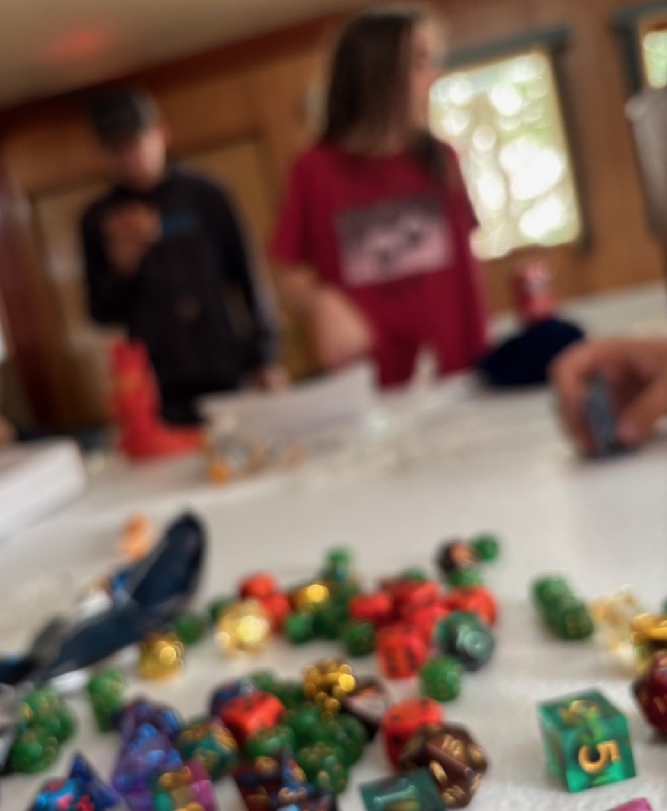 Colourful polyhedral dice scattered across a white table, with two people out of focus in the background.